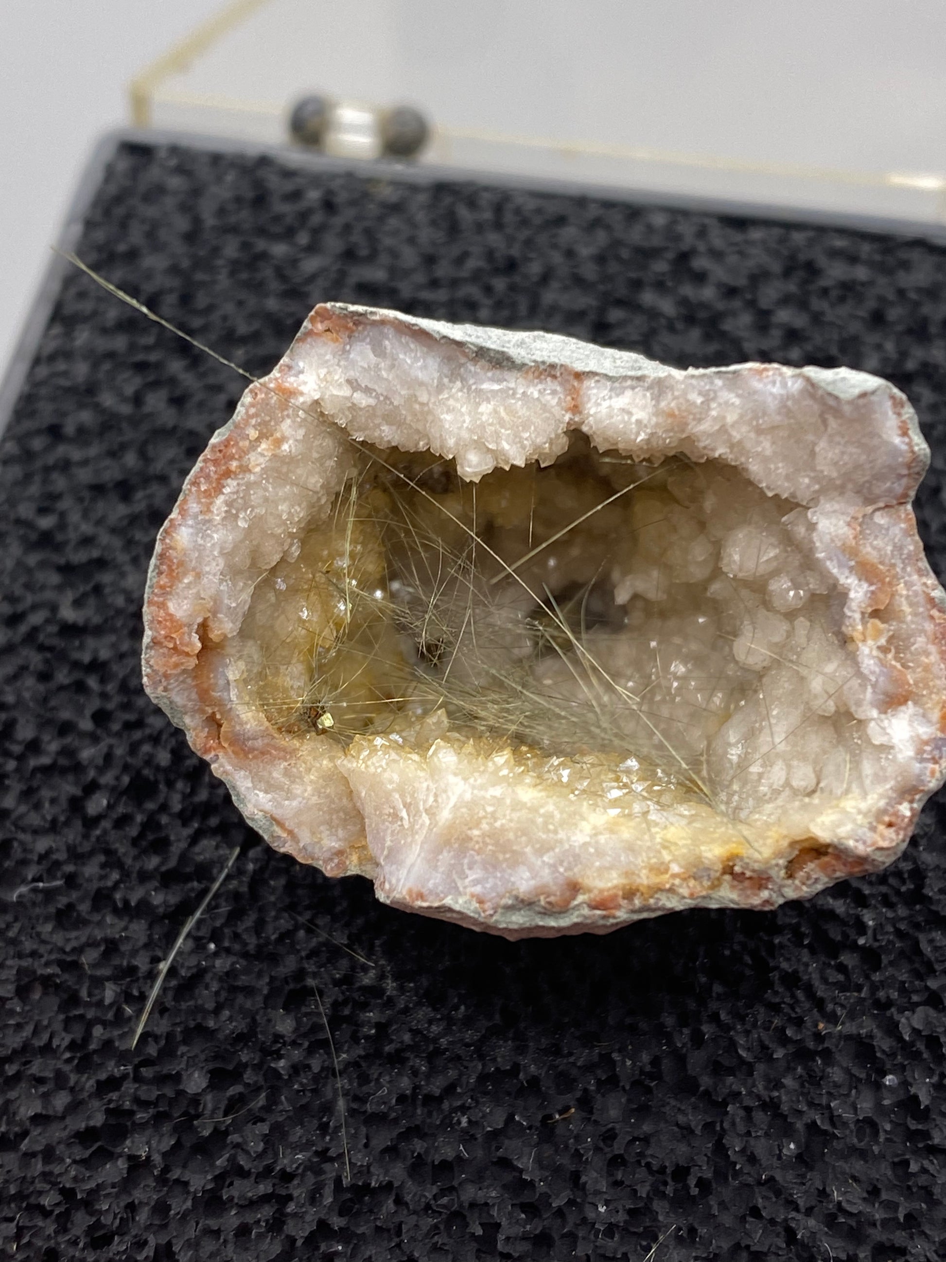 A halved geode filled with white and crystalline minerals, displaying thin, hair-like formations on the inside surface. Set on a textured black surface and likely part of a display, possibly in a museum or exhibit, it showcases the mesmerizing beauty found in The Crystalary's Millerite from Halls Gap, Lincoln Co., Kentucky.