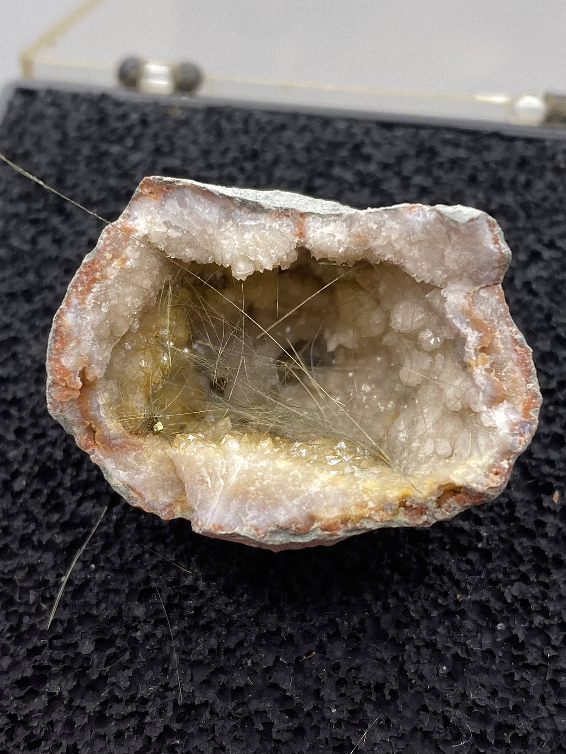 Close-up of The Crystalary's Millerite-Halls Gap geode from Lincoln Co., Kentucky, showcasing a rough outer surface and a hollow interior lined with white crystalline formations and golden-brown mineral deposits. Delicate, hair-like strands of Halls Gap Millerite extend across the cavity, embedded in a nickel-rich matrix. The geode rests on a black textured surface.