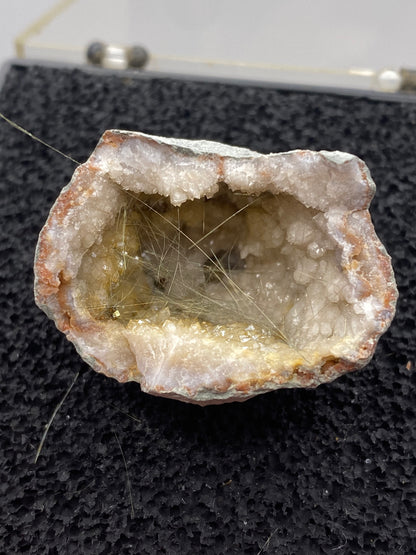 Close-up of The Crystalary's Millerite-Halls Gap geode from Lincoln Co., Kentucky, showcasing a rough outer surface and a hollow interior lined with white crystalline formations and golden-brown mineral deposits. Delicate, hair-like strands of Halls Gap Millerite extend across the cavity, embedded in a nickel-rich matrix. The geode rests on a black textured surface.