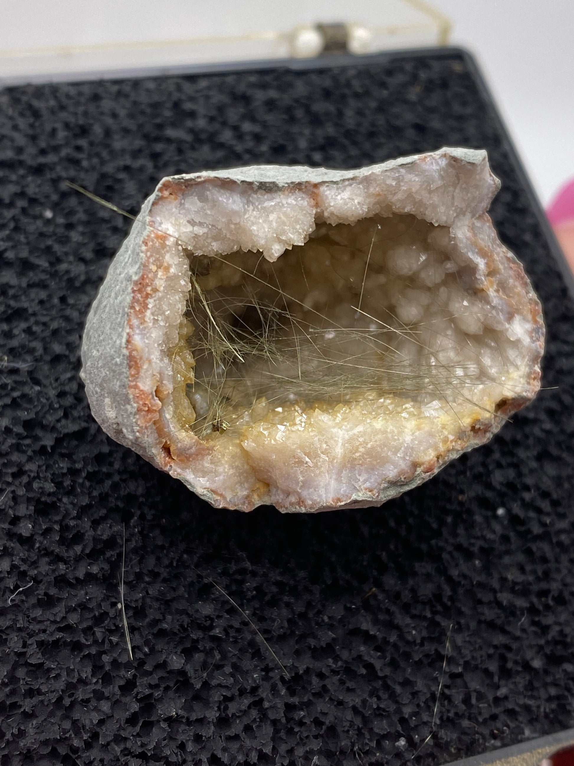 A close-up of a halved Millerite geode from Halls Gap, Lincoln Co., Kentucky, available from The Crystalary, reveals an interior lined with sparkling quartz crystals. Thin, needle-like crystals extend from the surface, hinting at the distinctive presence of Halls Gap Millerite. The geode rests on a textured black mat.