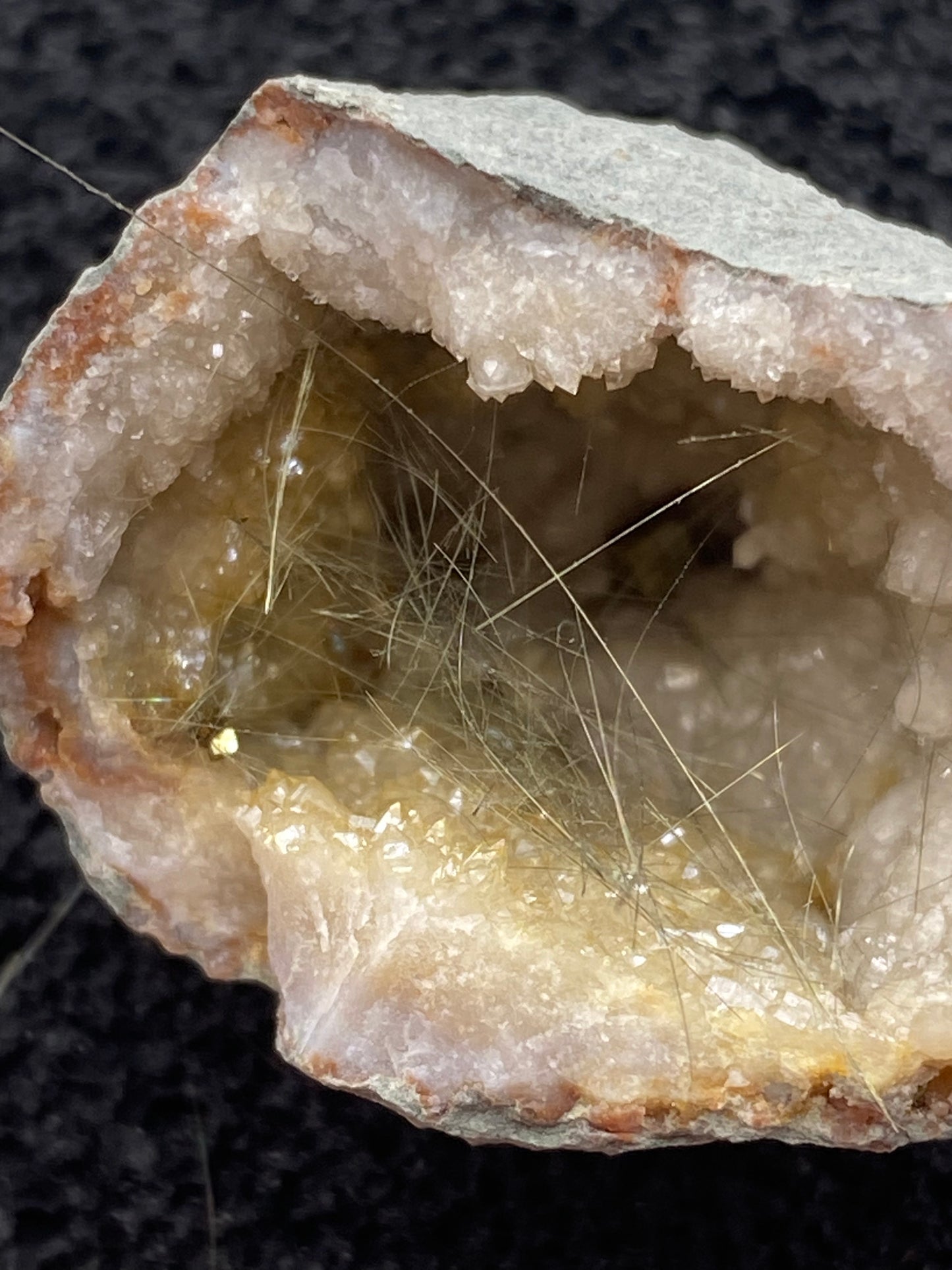 A close-up of a partially opened Millerite geode from Halls Gap, Lincoln Co., Kentucky, reveals intricate, thin needle-like crystals inside. The outer shell is rough and gray, contrasting with the sparkling, translucent interior minerals and delicate strands. This particular geode from The Crystalary features a nickel-rich matrix, adding to its charm as it rests on a dark surface.