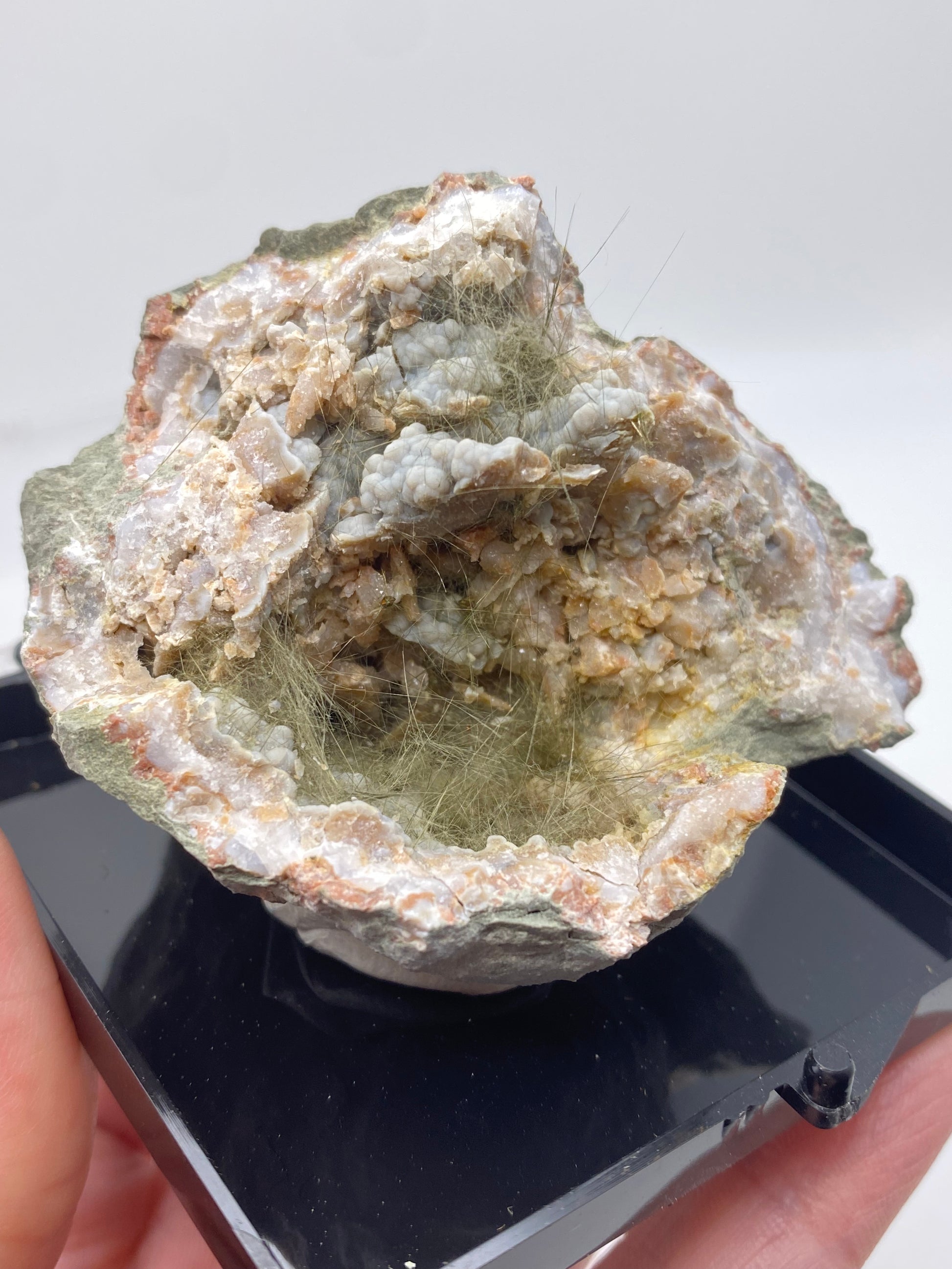 A person holds a chunk of rock displaying an exposed geode, likely one of the Millerite, Pyrite, Chalcedony pieces from Halls Gap, Lincoln Co., Kentucky. This prized specimen from The Crystalary features a variety of crystalline structures and delicate, hair-like mineral formations embedded within its cavity. The piece is placed on a black plastic base for support.