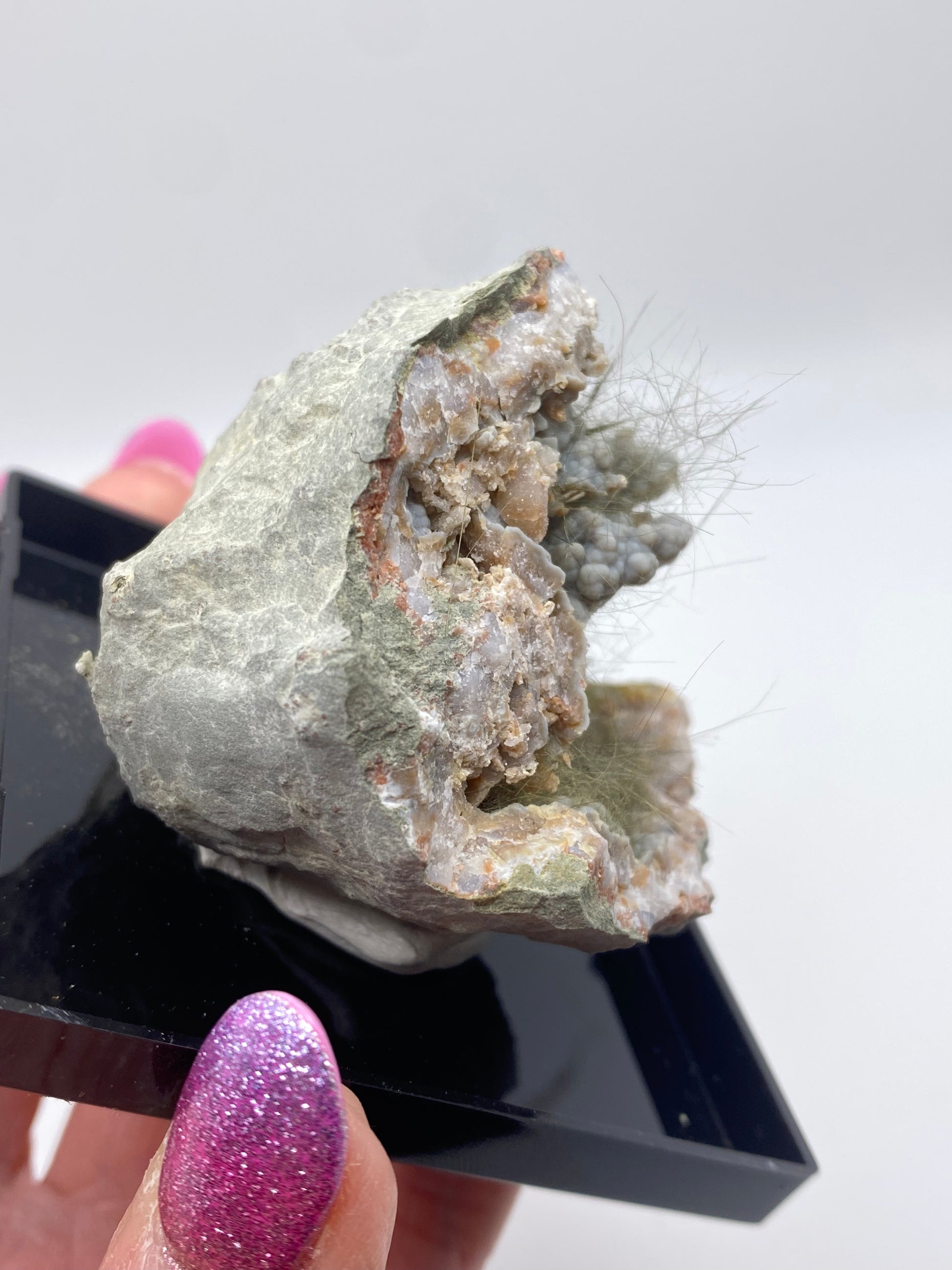 A person with glittery pink and purple nails holds a pale, rough rock specimen, identified as Millerite, Pyrite, Chalcedony from Halls Gap in Lincoln Co., Kentucky by The Crystalary, on a small black platform. The rock has a textured surface with jagged edges and some fibrous, hair-like strands protruding from its crevices.