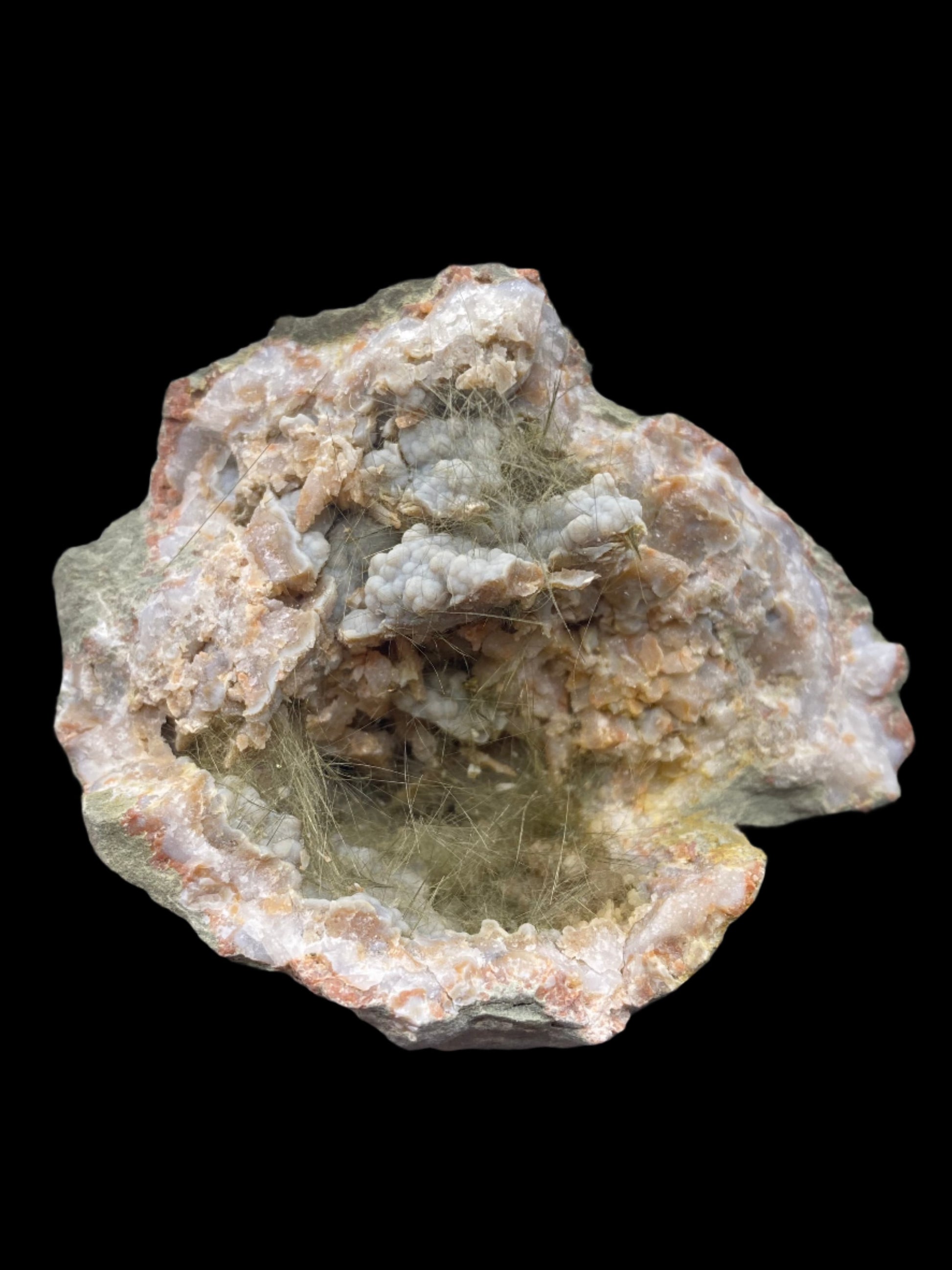 The Crystalary's Millerite, Pyrite, Chalcedony specimen from Halls Gap, Lincoln Co., Kentucky showcases a rough, irregular form with a mix of pale colors such as beige, light grey, and subtle hints of pink. Thin, fibrous strands are visible within the nickel-rich matrix, creating a textured effect against the solid black background.