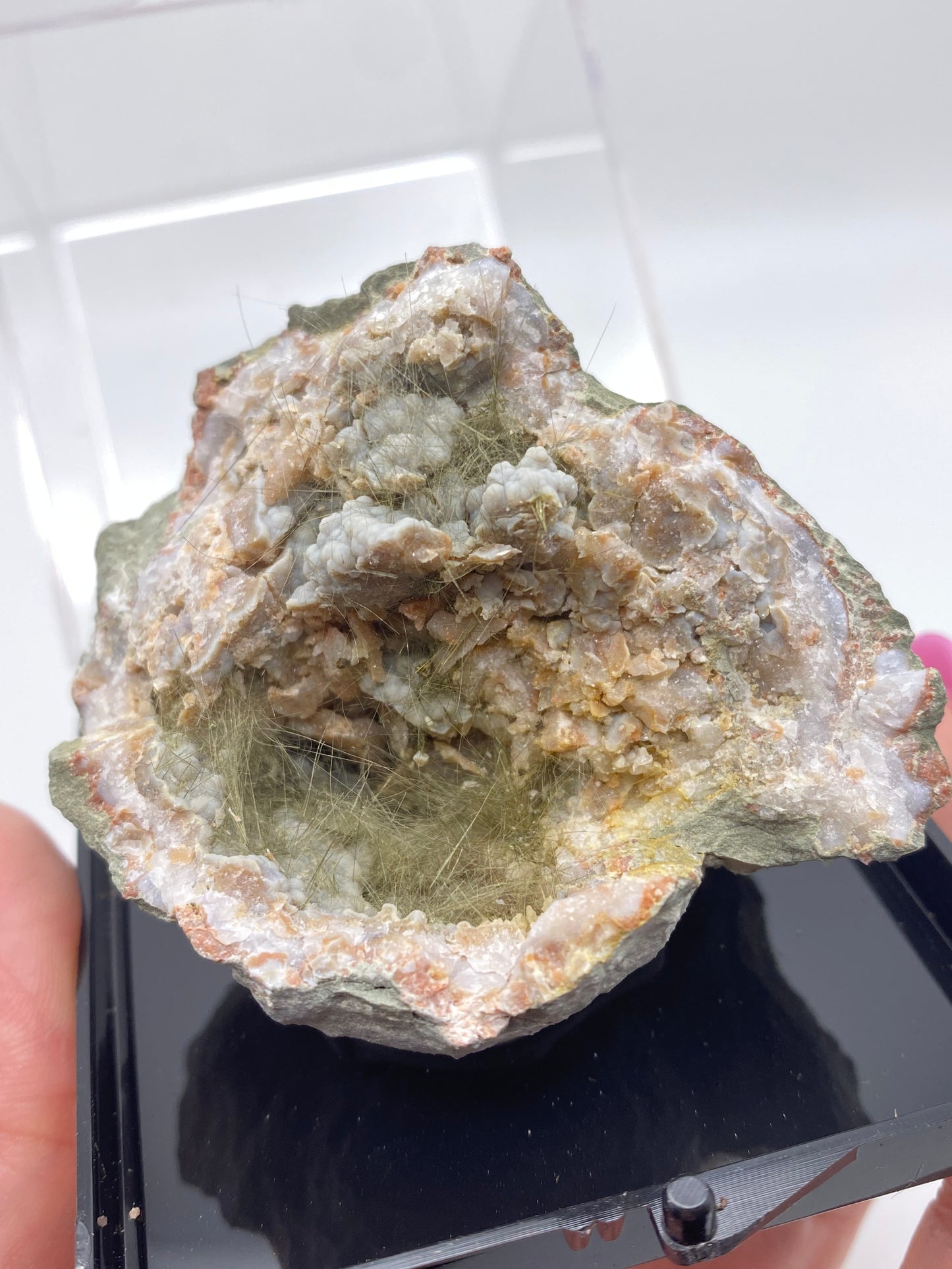 A rocky mineral specimen, which appears to be a Millerite, Pyrite, and Chalcedony from Halls Gap, Lincoln Co., Kentucky by The Crystalary, is displayed on a black stand. The mineral has a rough, uneven surface with a mix of beige and pale green colors. Thin, hair-like strands are visible within some parts of the rock's nickel-rich matrix. The background is white, and the rock is in focus.