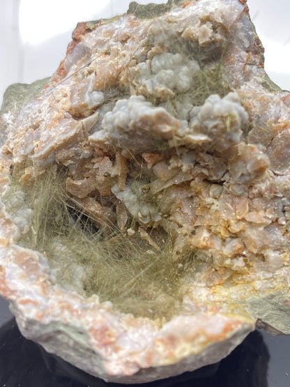 A close-up of a cut Kentucky geode from The Crystalary showcases an interior filled with light-colored chalcedony crystals and a network of delicate, greenish millerite fibrous formations. The nickel-rich matrix contrasts with the rough and weathered exterior, highlighting the intricate structures inside.