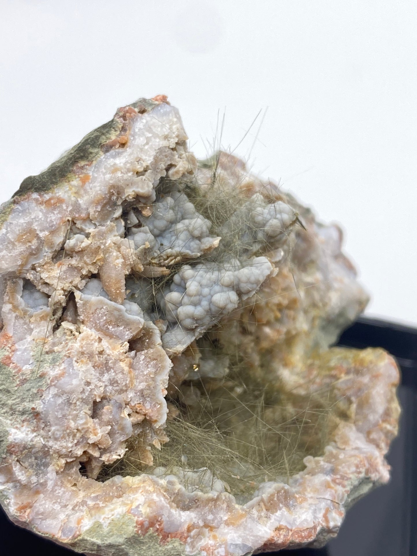 A close-up of The Crystalary's Millerite, Pyrite, Chalcedony specimen from Halls Gap, Lincoln Co., Kentucky showcases a rough rock exterior that reveals a mesmerizing cluster of light green, hair-like crystals accompanied by bluish-gray stone formations inside. This captivating Kentucky geode is elegantly displayed on a black surface against a white background.