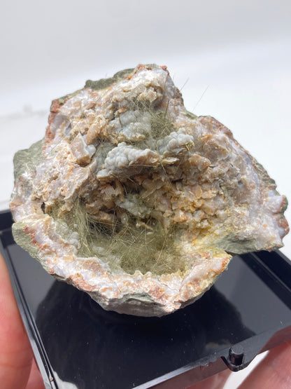 A close-up of a hand holding a mineral specimen from The Crystalary's collection reveals fibrous inclusions within a rugged, rocky matrix. The slender, light-colored fibers stand out against the surrounding grayish and brownish rock typical of Kentucky geodes. This particular specimen features Millerite, Pyrite, and Chalcedony from Halls Gap, Lincoln Co., Kentucky, and is placed on a black surface.