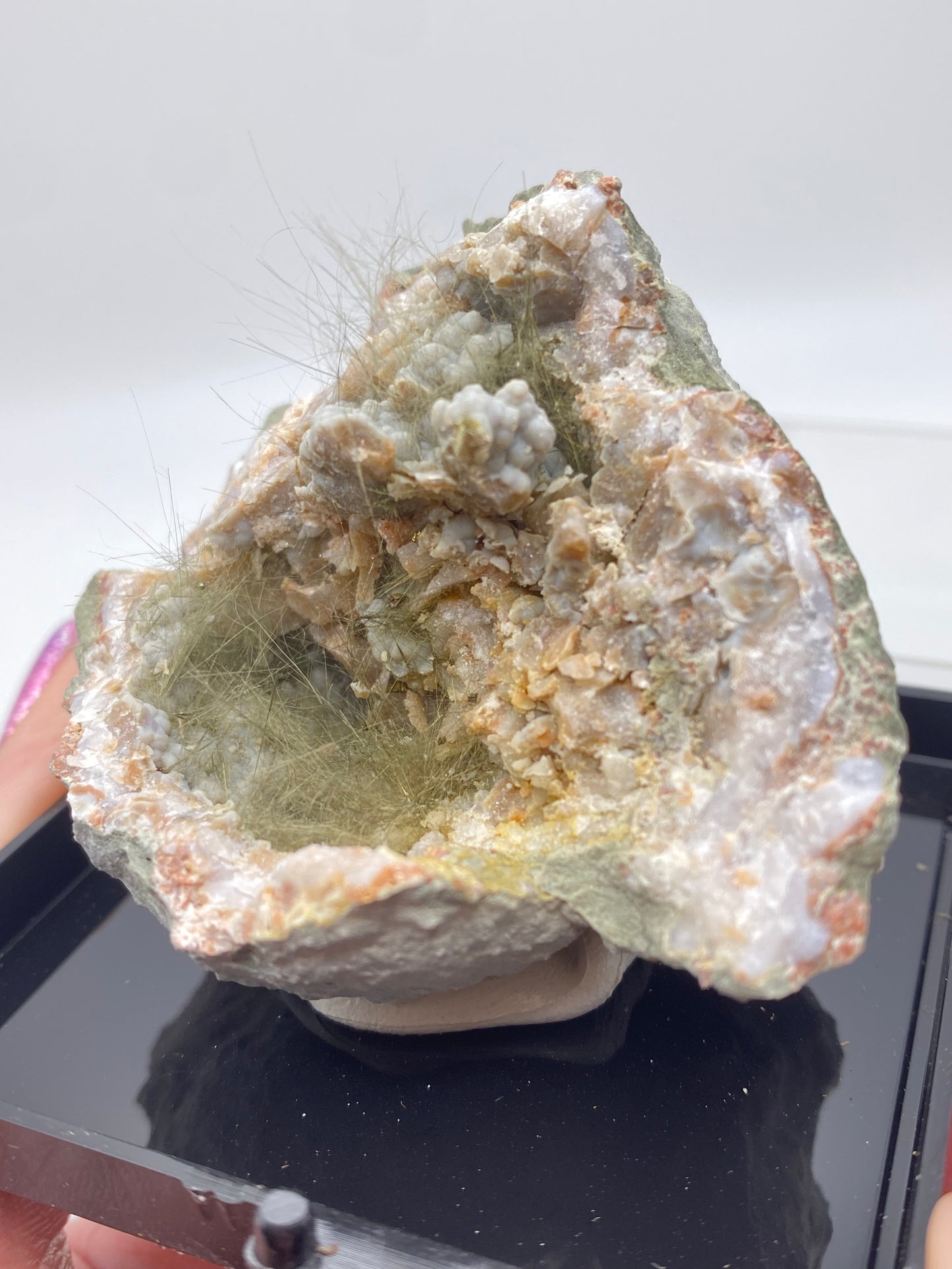 A close-up photo of a Millerite, Pyrite, and Chalcedony specimen on display. The rock, sourced from Halls Gap, Lincoln Co., Kentucky by The Crystalary, has a rough, uneven surface with greenish hues and contains fibrous, hair-like crystal formations inside. The specimen is placed on a black tray against a plain white background.