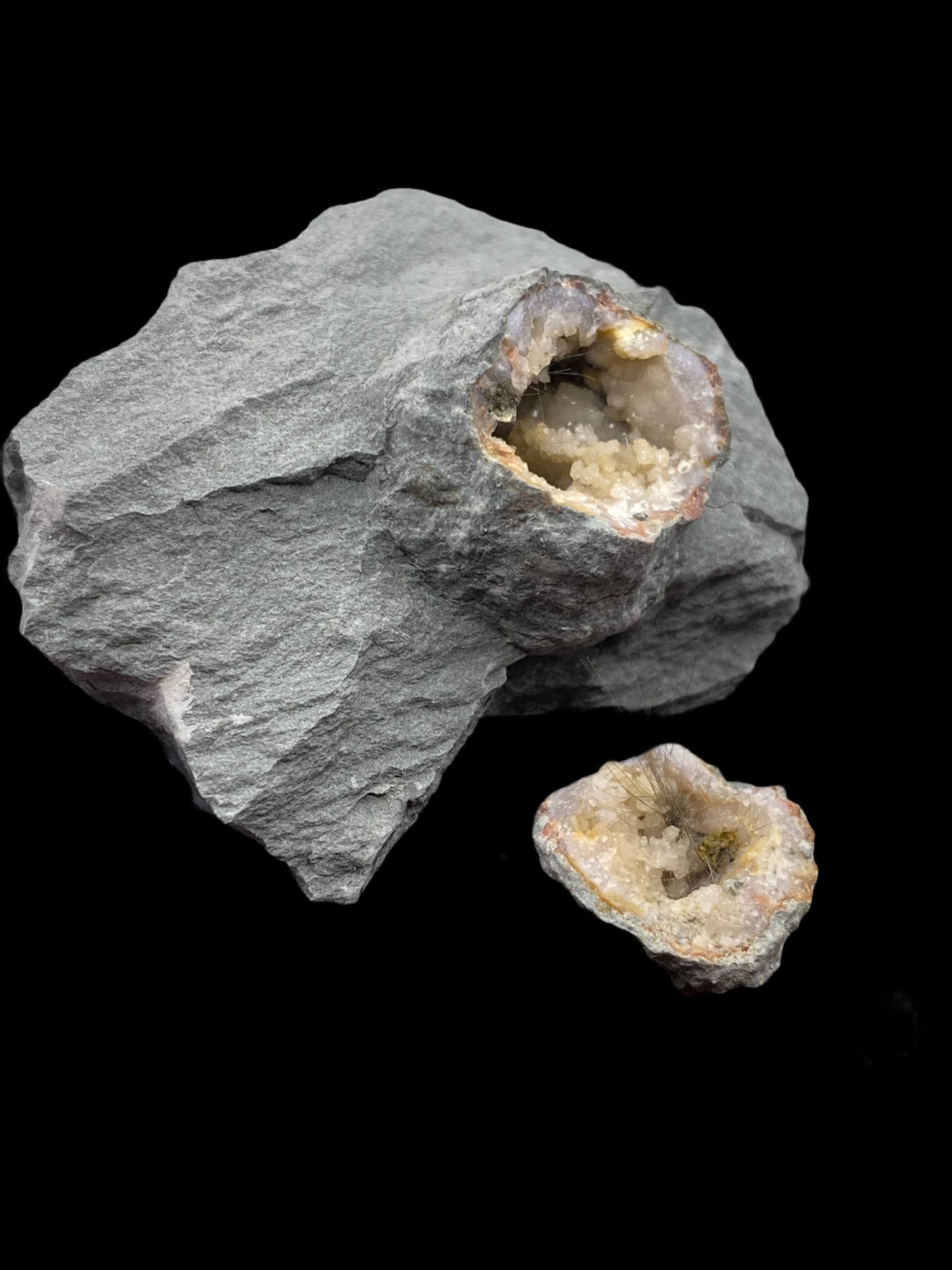 Two rocks from "The Crystalary's" Millerite, Quartz collection from Halls Gap, Lincoln Co., Kentucky rest against a black background. The larger rock features a visible geode cavity filled with crystalline formations, while a smaller geode sits in front, also showcasing its inner crystals. The light-hued quartz pyrite calcite crystals contrast beautifully with the grey outer rock, creating a striking visual effect.