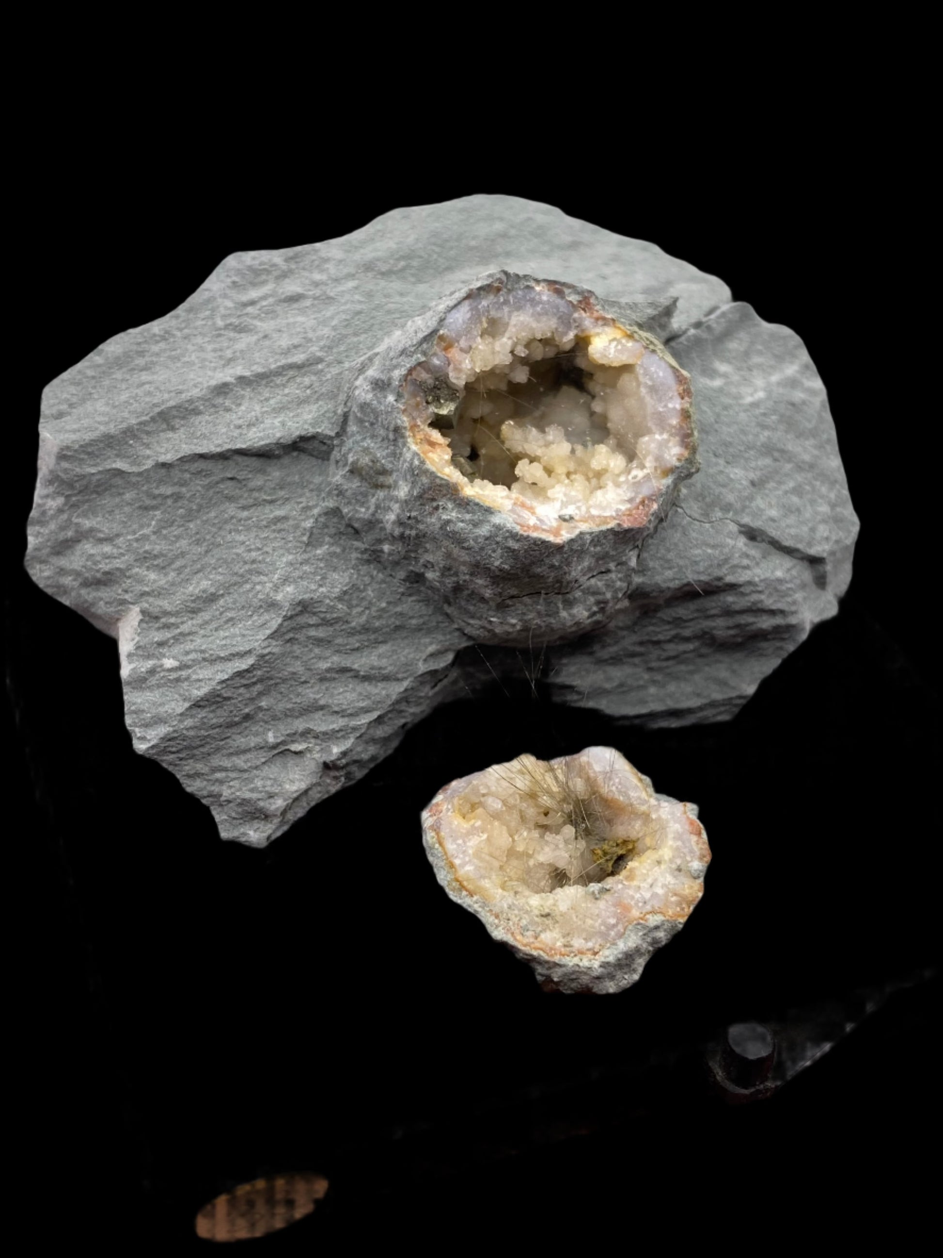 Displayed against a black background, an impressive quartz pyrite specimen from The Crystalary showcases a rough gray rock with a hollow cavity filled with small, sparkling crystals. Below it lies another smaller piece of rock featuring a similar glittering crystal-filled cavity, both originating from Halls Gap, Lincoln Co., Kentucky.