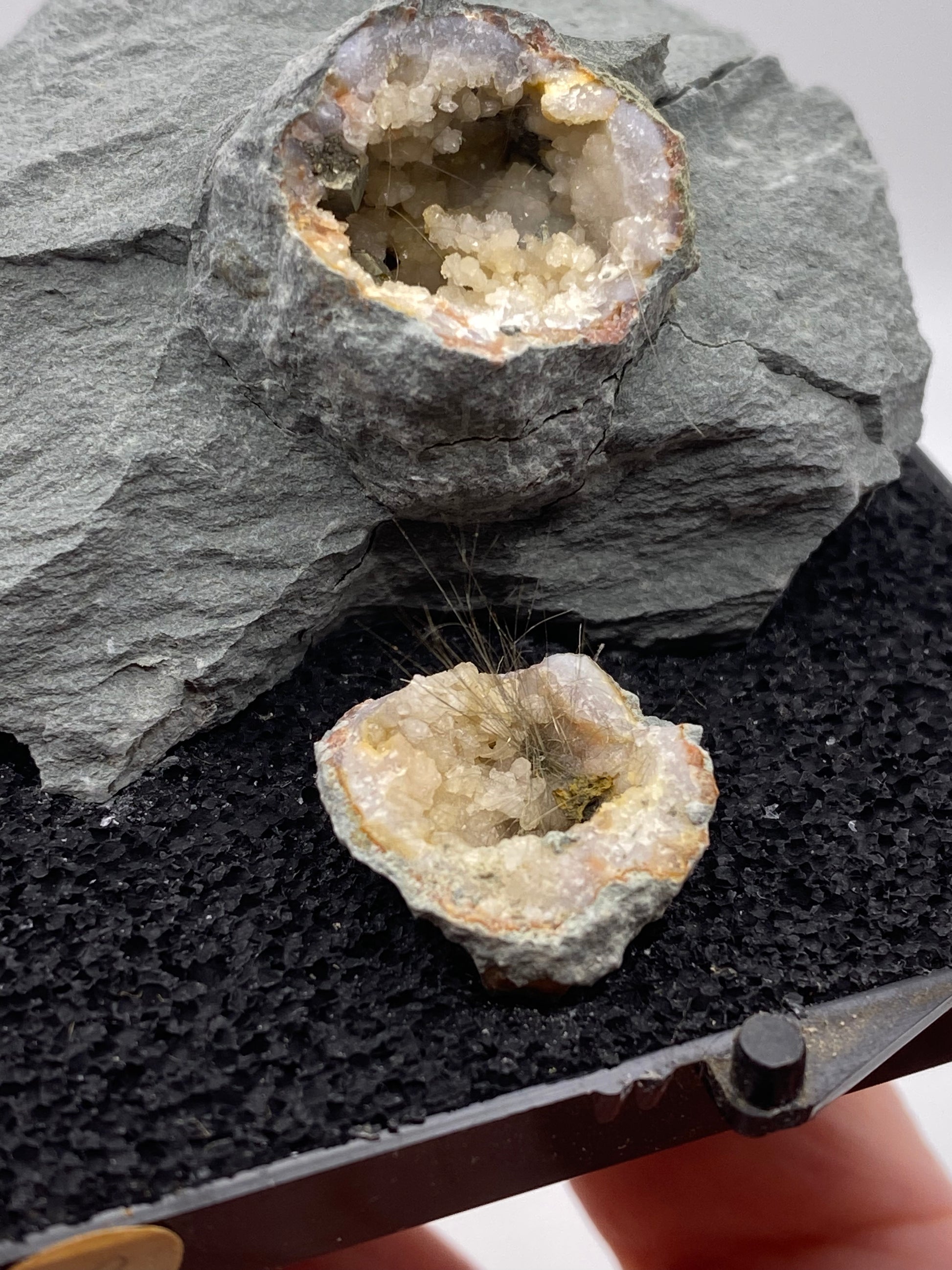 Close-up of two geodes, hollow mineral formations with crystals inside, placed on a rough gray stone. The top geode is filled with small, white crystals, while the bottom one from "The Crystalary" showcases a stunning Millerite and Quartz combination from Halls Gap, Lincoln Co., Kentucky. It features white crystals interspersed with fine hair-like minerals amidst a nickel-rich matrix.