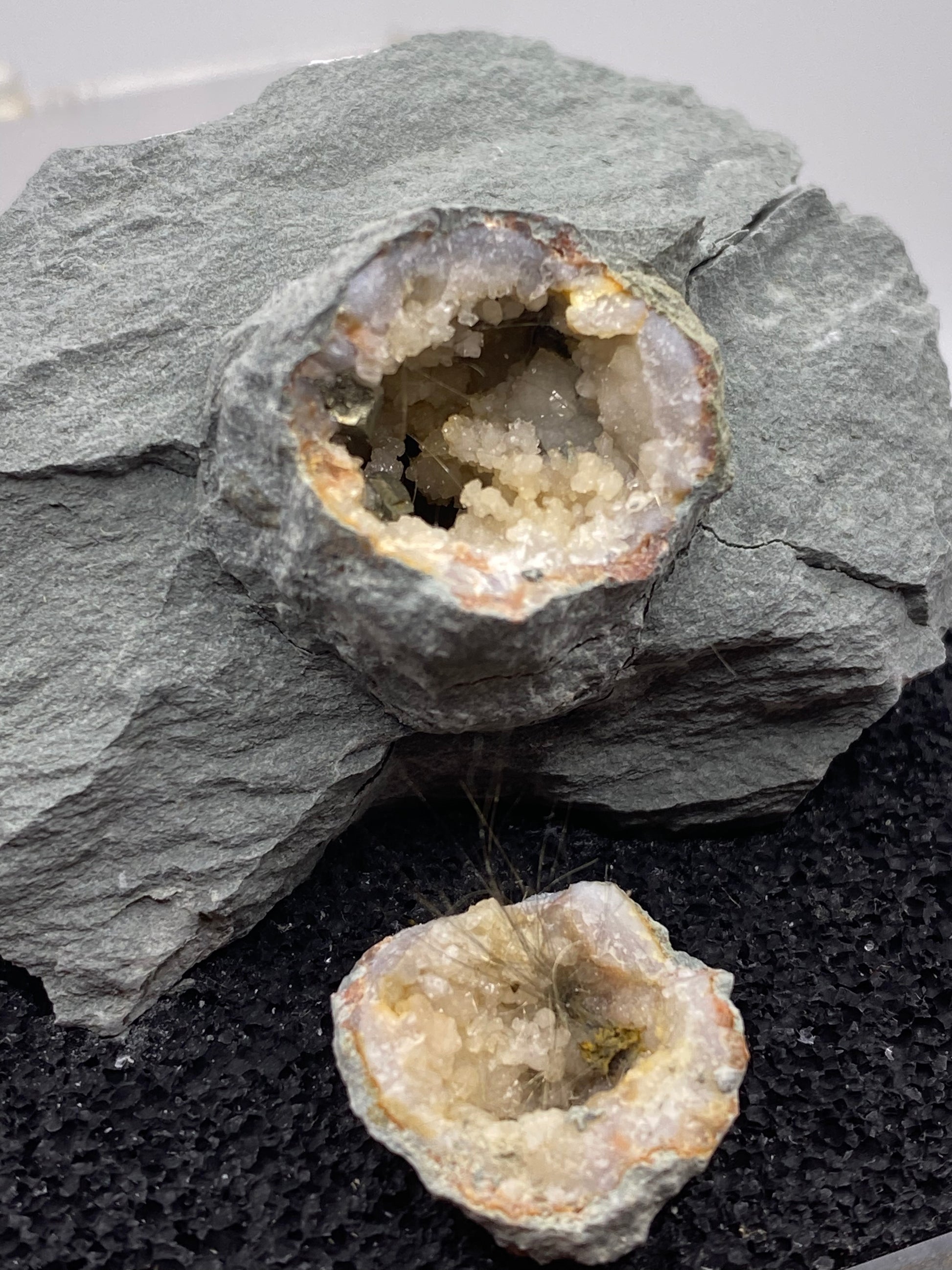 A split Millerite geode from The Crystalary rests on a rough, gray stone surface, revealing its interior filled with sparkling, white, and clear crystalline formations. The top half of the geode is placed on top of the stone, showcasing a nickel-rich matrix speckled with hints of quartz and pyrite from Halls Gap, Lincoln Co., Kentucky.