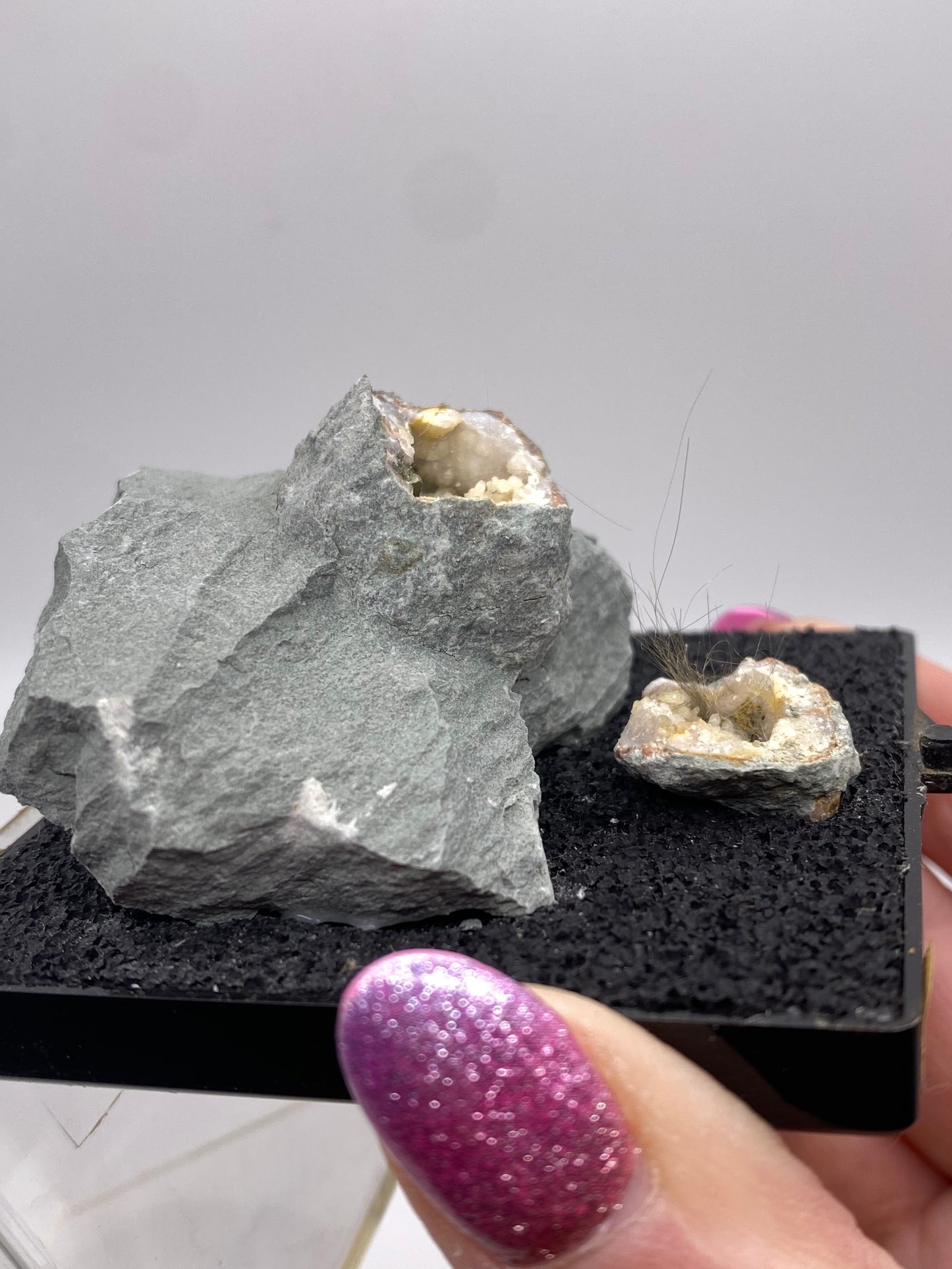 A hand with sparkly purple nails holds a small black stand that displays two rough rocks, one larger and one smaller. Both rocks have cavities lined with crystalline formations, showcasing the unique beauty of Millerite and Quartz geodes from Halls Gap, Lincoln Co., Kentucky. The background is plain and out of focus. Product by The Crystalary.