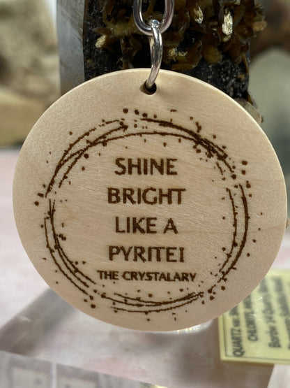 Introducing the Mineral/Crystal Theme Keychain by The Crystalary: a wooden keychain featuring intricate circular laser engraving that reads "SHINE BRIGHT LIKE A PYRITE! THE CRYSTALARY." The text is surrounded by an elegant circular design, making it one of the most captivating mineral-themed keychains. The keychain also includes a sturdy metal ring for easy hanging.