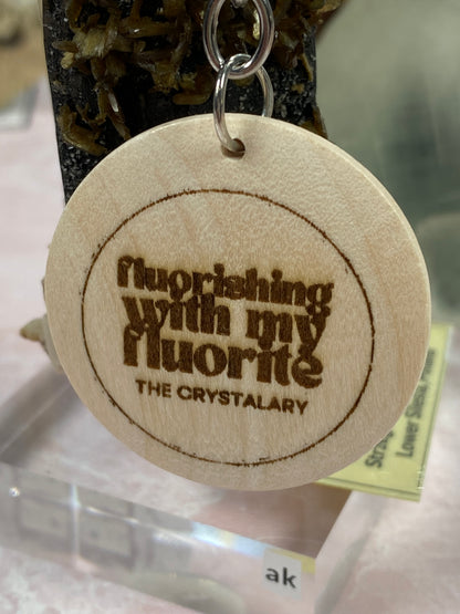 Introducing the Mineral/Crystal Theme Keychain from The Crystalary: a round, wooden keychain laser-engraved with "flourishing with my fluorite" and "THE CRYSTALARY." This charming accessory, attached to a metal ring, perfectly captures the allure of mineral-themed keychains. In the background, a display stand is subtly visible.