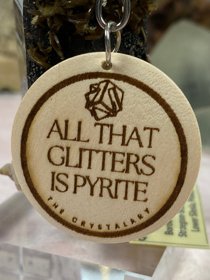 This laser engraved keychain from The Crystalary, part of their Mineral/Crystal Theme Keychains collection, features a round wooden base with a brown engraving that reads "All That Glitters Is Pyrite." Above the text is a striking geometric design. Displayed against a rock with mineral formations in the background, it’s an ideal accessory for anyone who loves mineral-themed keychains.