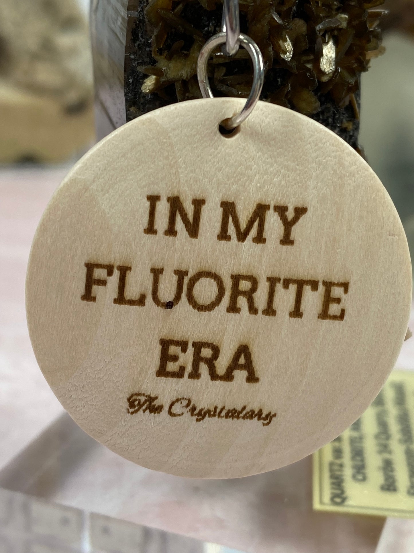 A circular wooden keychain, laser engraved with "IN MY FLUORITE ERA" and "The Crystalary," hangs elegantly from a metal ring. The background features a stunning piece of fluorite mineral on display, with a softly blurred informational card at the bottom. This keychain is part of the Mineral/Crystal Theme Keychains collection by The Crystalary.