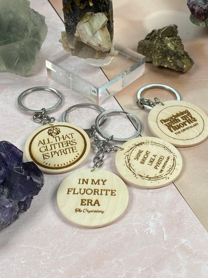 A collection of The Crystalary's Mineral/Crystal Theme Keychains, featuring four wooden pieces with laser-engraved text, displayed on a pink surface among mineral and crystal specimens. The keychains read: "ALL THAT GLITTERS IS PYRITE," "IN MY FLUORITE ERA," "SHINE BRIGHT LIKE A PYRITE," and "LESSON IN RUTILE.