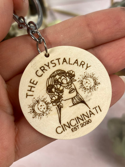 A close-up of a hand holding a wooden keychain from The Crystalary's Mineral/Crystal Theme Keychains collection. The keychain features an engraved image of a woman wearing a flower crown, surrounded by the text "THE CRYSTALARY CINCINNATI EST 2020." Adorned with small moon and star illustrations, this laser-engraved design is perfect for those who love mineral-themed accessories.