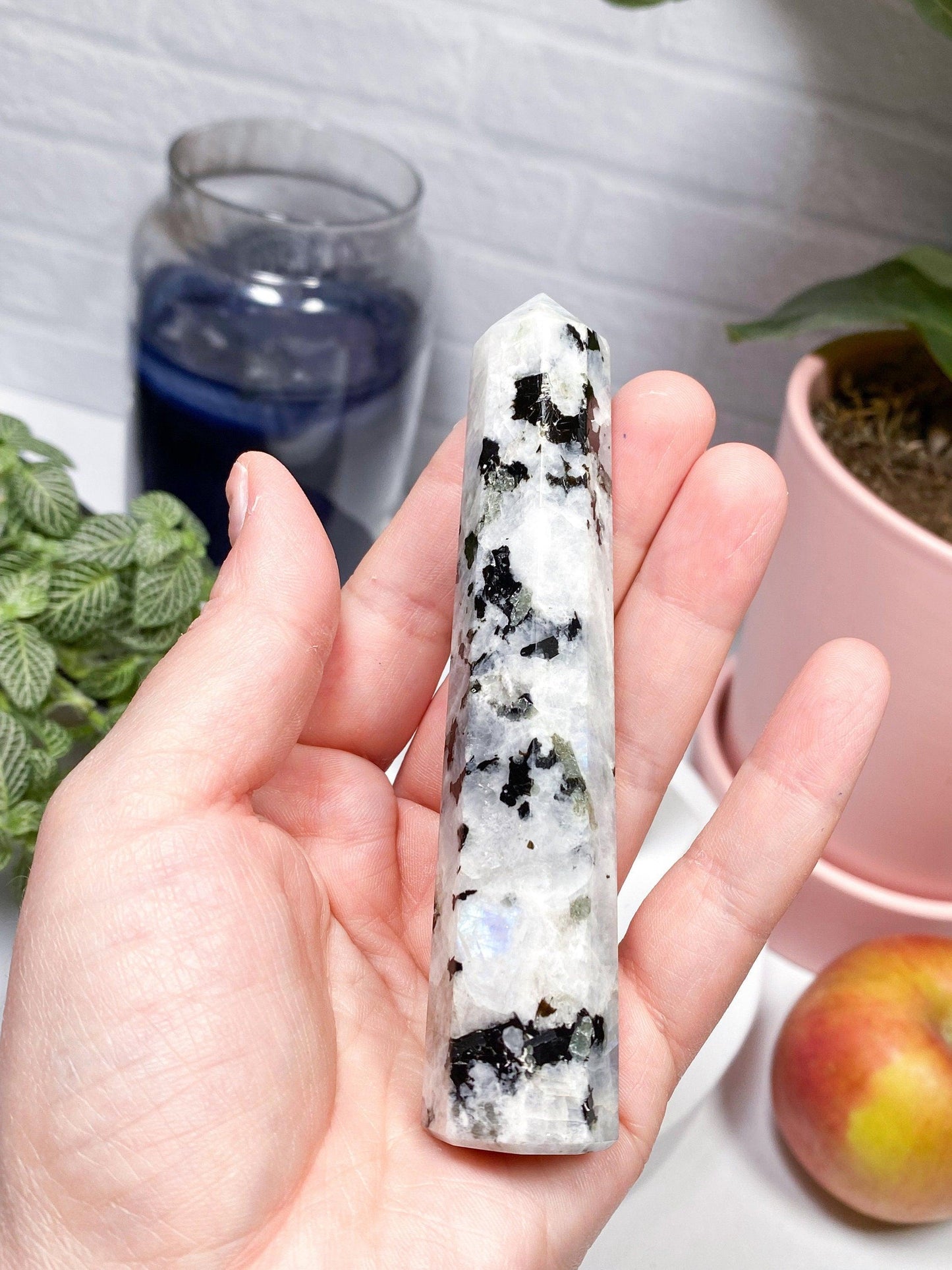 A hand holds a polished, pointed Moonstone Crystal Point from The Crystalary, showcasing its black inclusions. In the background, a blue candle, green plant, pink pot, and an apple rest on a white surface. This intuitively chosen gem features 8-sided points and evokes serene beauty in a well-lit room with a white brick wall.