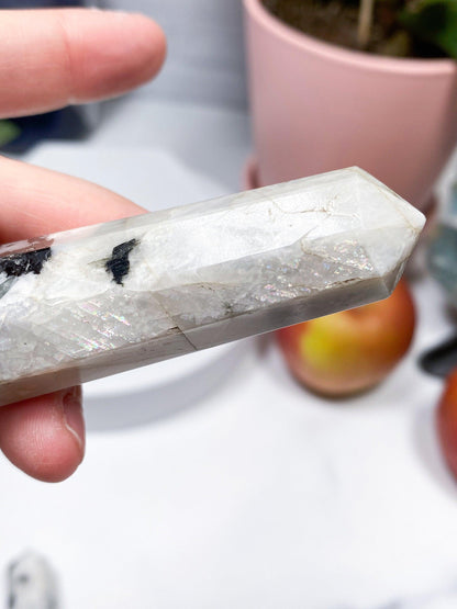 A hand holds a polished Moonstone Crystal Point from The Crystalary, showcasing its 8-sided formation, white and slightly translucent appearance, and unique black streaks within. In the background, there is a pink plant pot, some partially blurry fruit, and a gentle, well-lit indoor setting.