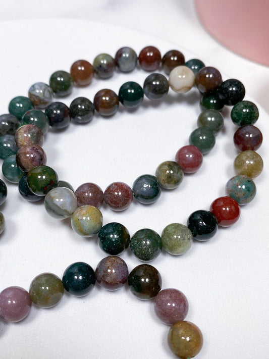 A close-up view of a beaded gemstone necklace, reminiscent of The Crystalary’s Moss Agate Crystal Bracelet. The round beads come in various colors including shades of green, brown, red, purple, and white. The necklace is coiled in a loose spiral pattern on a white surface with a hint of a pink object in the background.