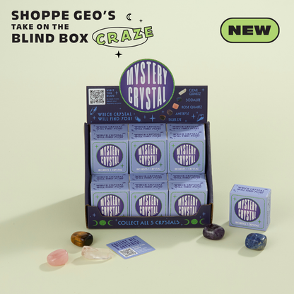 A display box labeled "Mystery Crystal Blind Box Assortment - Stocking Stuffers!" by GeoCentral contains individual packages of crystals. Four loose crystals of various colors are displayed in front. Text reads "SHOPPE GEO'S TAKE ON THE BLIND BOX CRAZE" and "NEW". The light green background makes this Crystal Collection a must-have for enthusiasts.