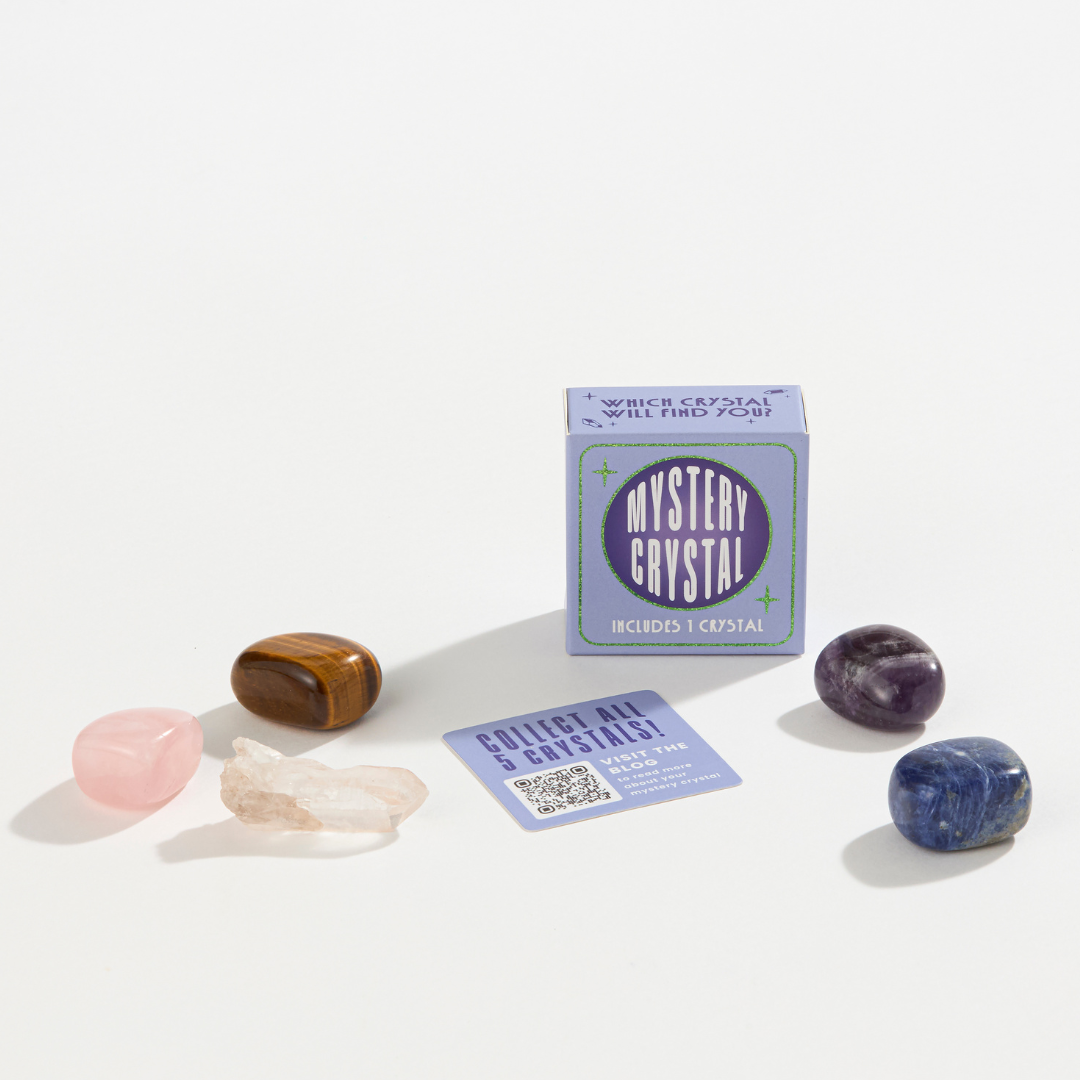 A small, light purple box labeled "Mystery Crystal Blind Box Assortment - Stocking Stuffers!" by GeoCentral is surrounded by five polished crystals in various colors: pink, brown, white, purple, and blue. Beside the box, an informational card encourages the collection of all five crystals in this delightful assortment.