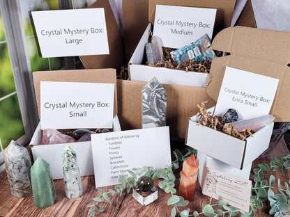 A display featuring The Crystalary's "Mystery Crystal Box – Extremely High Quality" in sizes Extra Large, Large, Medium, and Small. Each intricately designed mystery box contains premium crystal items. Surrounding the boxes are numerous standalone polished crystals and greenery, accompanied by informational cards detailing the contents.