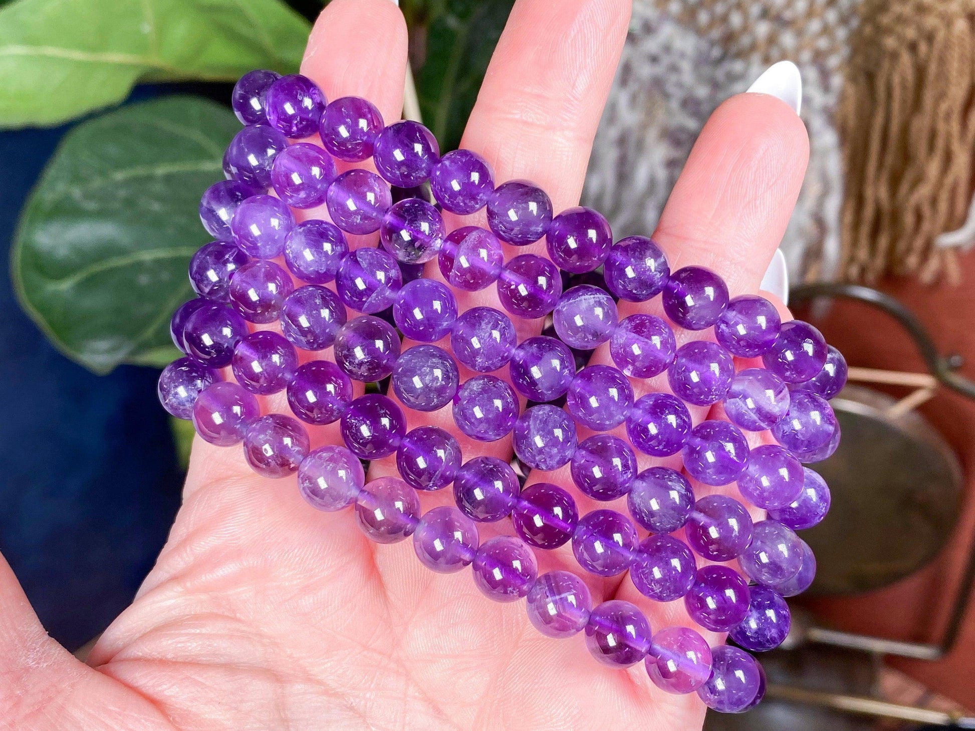 A hand gently holds several strands of round, polished amethyst beads from The Crystalary's Natural Amethyst Crystal Bracelet, showcasing their vibrant purple hues. The background features a blurred green plant and a mix of earthy tones, emphasizing the natural gemmy Amethyst's reputed anxiety relief properties.