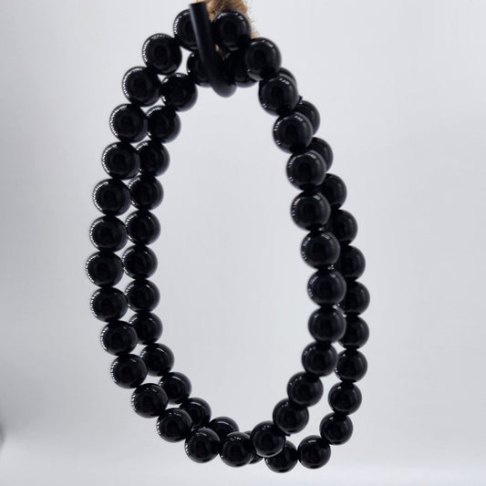 An Obsidian Crystal Bracelet, renowned for its protective properties against negative energy, is displayed hanging on a twine-wrapped circular object by The Crystalary. The plain, light-colored background accentuates both the bracelet and the twine-wrapped object.