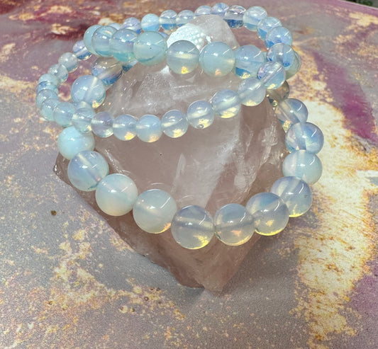 A close-up of three Opalite Crystal Bracelets by The Crystalary, known for their mood stabilization properties, draped over a large, rough-cut, translucent ethical crystal. The crystal and bracelets are displayed on a textured surface with shades of rust, pink, and gray. The opalite beads are iridescent, reflecting a soft, luminous glow.