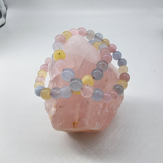 The Pastel Crystal Bracelet by The Crystalary is draped over a large pink quartz crystal. The bracelet, featuring smooth round beads in delicate shades of pink, blue, and yellow, contrasts beautifully with the rough texture of the quartz. This charming accessory rests on a plain white surface, emphasizing its soft pastel hues.