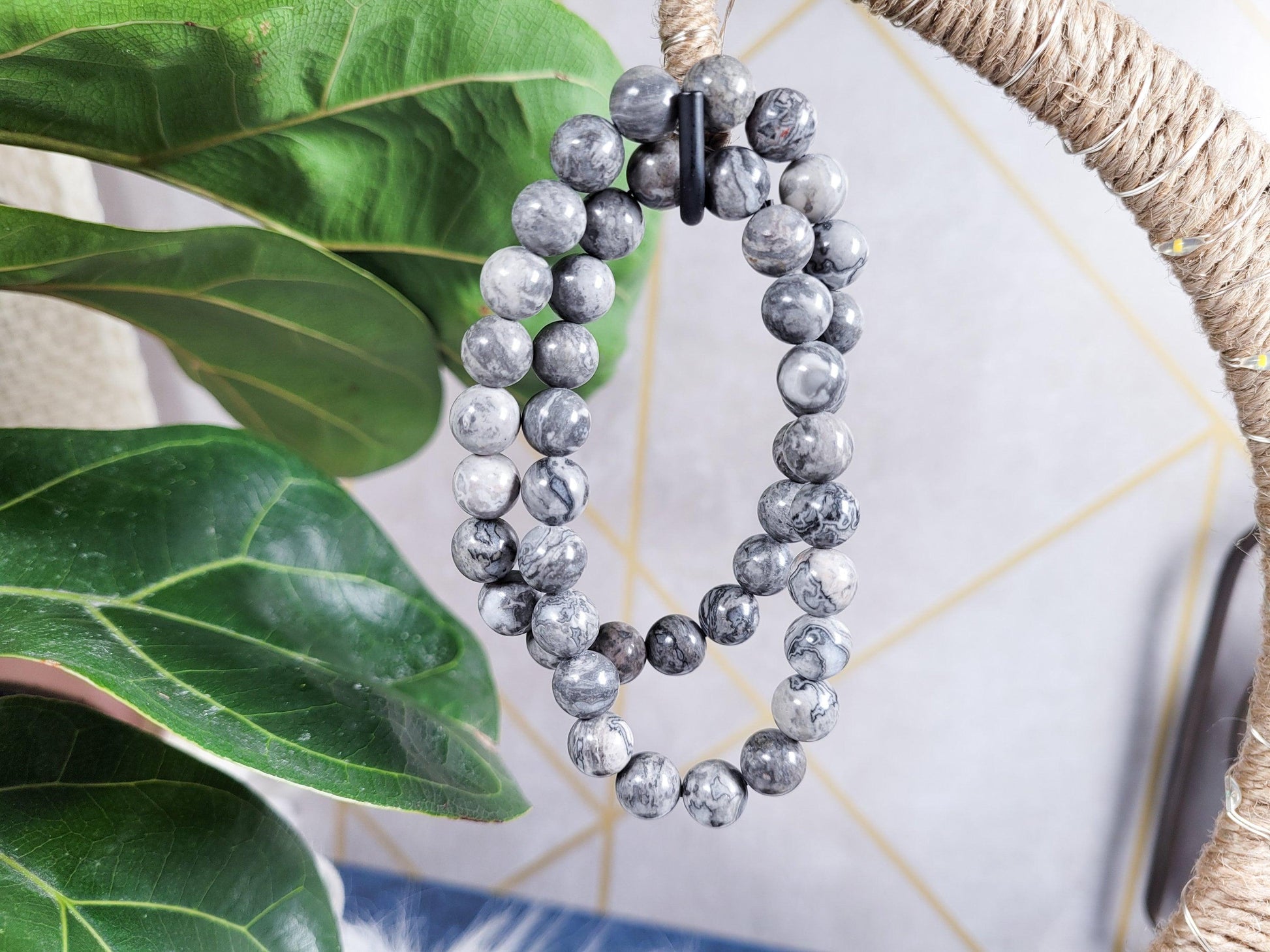 A set of two Picasso Jasper Crystal Bracelets from The Crystalary, featuring marbled gray and black patterns, hangs on a wrapped cord. The background showcases lush green leaves and a light-colored tiled wall with geometric patterns, adding an elegant touch to these gemstone bracelets sourced from the ethical crystal trade.