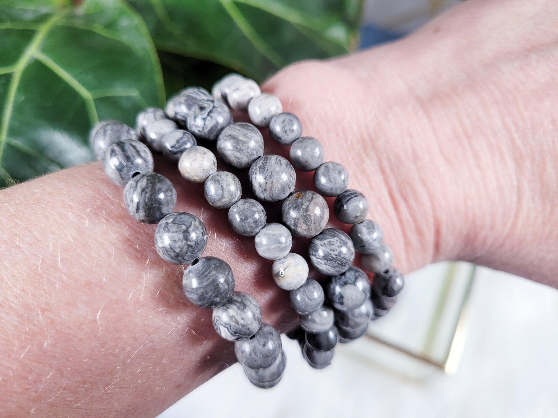 A close-up of a wrist adorned with multiple strands of round gray and white marbled stone beads, including Picasso Jasper Crystal Bracelets by The Crystalary. The background features green leaves, adding a subtle touch of nature to the image.