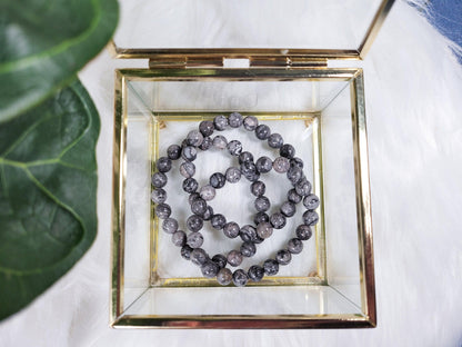 A gold-framed glass box from The Crystalary holds a necklace and a bracelet made of grey marbled beads. The box, resting on a white fluffy surface with a green leaf partially visible in the corner, showcases the exquisite Picasso Jasper crystal bracelets. The polished beads create an elegant and luxurious presentation.