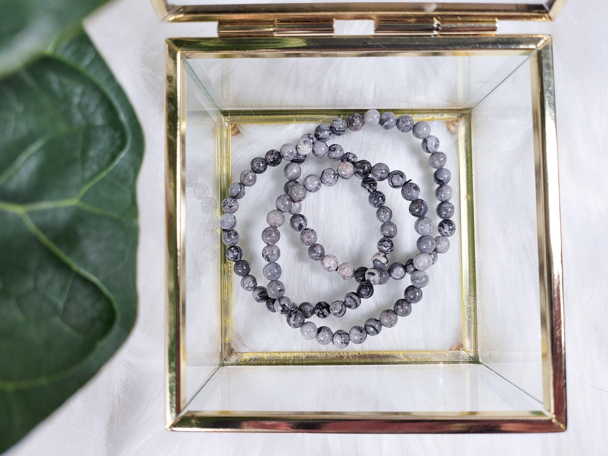 A glass and gold jewelry box containing three gray Picasso Jasper crystal bracelets from The Crystalary is placed on a white, fluffy surface, with a green leaf partially visible on the left side of the image. These bracelets are sourced through an ethical crystal trade, ensuring their beauty is matched by responsible practices.