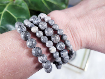 A wrist adorned with several grey and black marbled beaded bracelets. The beads vary slightly in color and pattern, creating a harmonious, natural appearance. Among them are Picasso Jasper Crystal Bracelets from The Crystalary, sourced through ethical crystal trade. The background features green foliage.