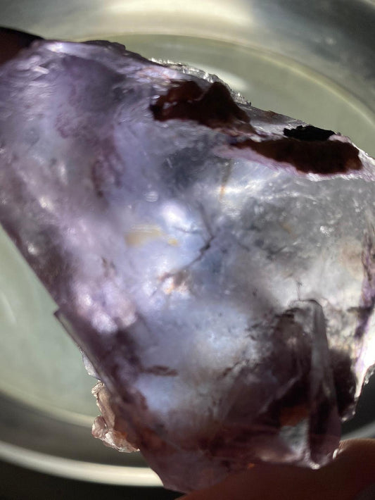 Close-up view of a Private Vault Session mineral by The Crystalary, showcasing its rough, unpolished beauty with a translucent purple hue. Held partially against the light, the image emphasizes its intricate internal structure and texture. With an out-of-focus background, this stunning mineral takes center stage—a true gem among specimens.