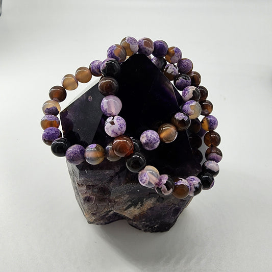 A polished amethyst crystal cluster from The Crystalary serves as a display stand for two Purple Agate Crystal Bracelets, each strung on elastic bands with round, multicolored stones blending shades of purple, brown, black, and white. The plain white background highlights the protective crystal and bracelets beautifully.