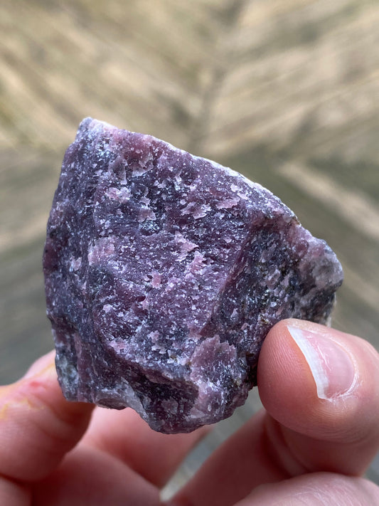 A hand holds a rough, unpolished purple Purpurite rock with specks of white and gray minerals. This raw specimen from The Crystalary’s Purpurite collection, sourced from Maine, weighs 105 grams. The background is blurred, emphasizing a textured surface with natural colors.