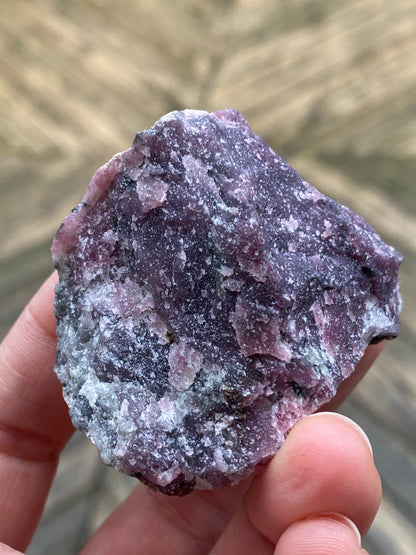 A hand is holding a rough, unpolished mineral specimen of raw Purpurite, Maine from The Crystalary, weighing 105 grams. Predominantly purple and white with visible crystalline textures, this gem hails from Maine. The background is out of focus, highlighting the intricate details and vibrant colors of the mineral.