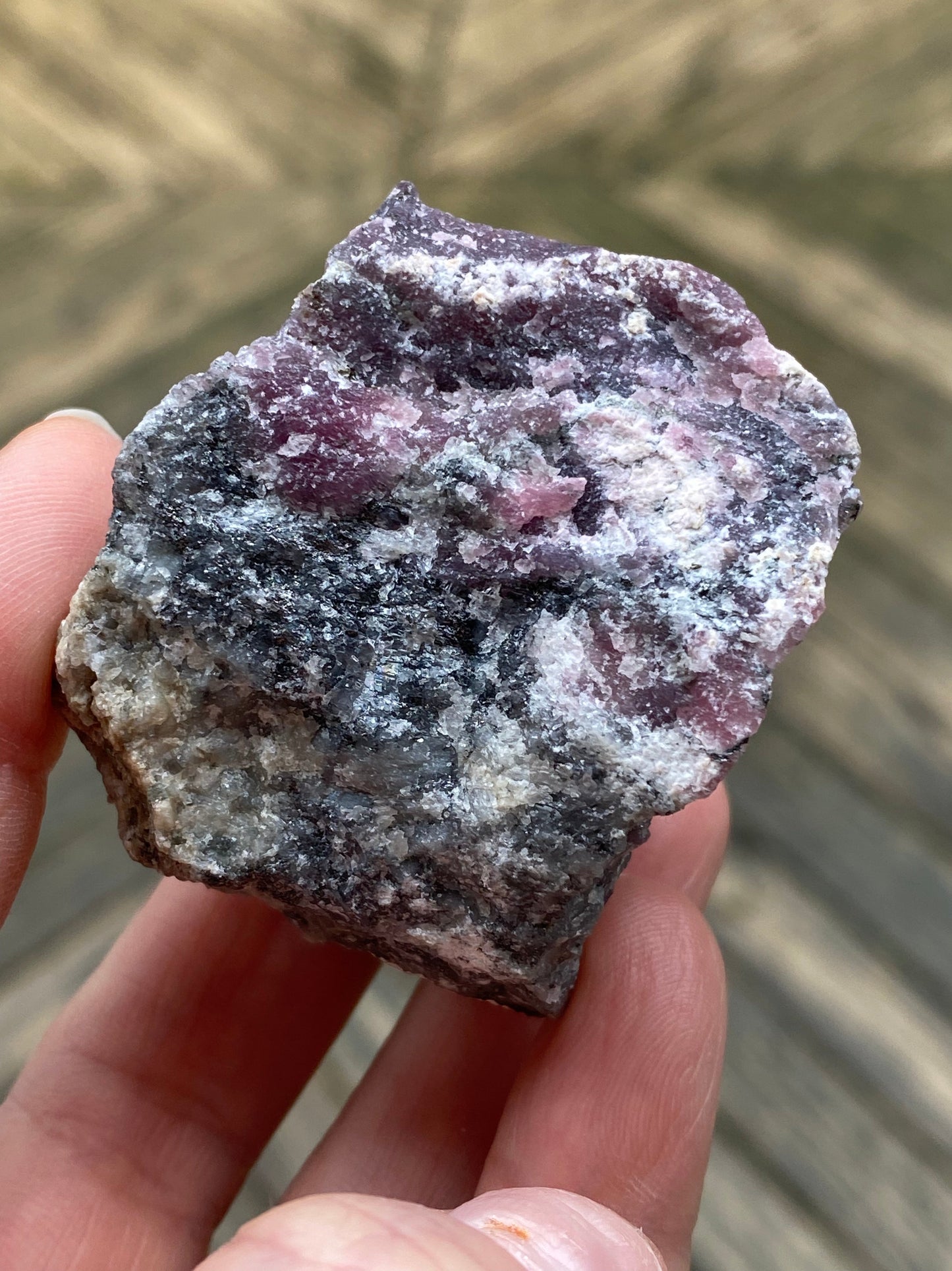 A close-up photo of a hand holding a 105-gram raw specimen of "Purpurite, Maine" by The Crystalary. This mineral, sourced from Maine, exhibits a blend of purple, black, and white hues with a coarse texture and uneven surface, captured against a blurred background.