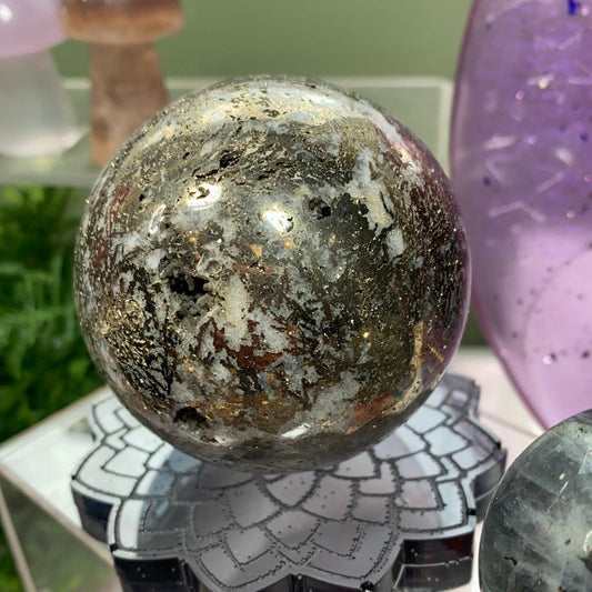 A Pyrite in Quartz Sphere from The Crystalary, featuring a metallic, marbled appearance, is displayed on an intricate, flower-shaped stand. Surrounding the sphere are various other crystals and decorative items, including one with etched purple details and another piece of glimmering pyrite.