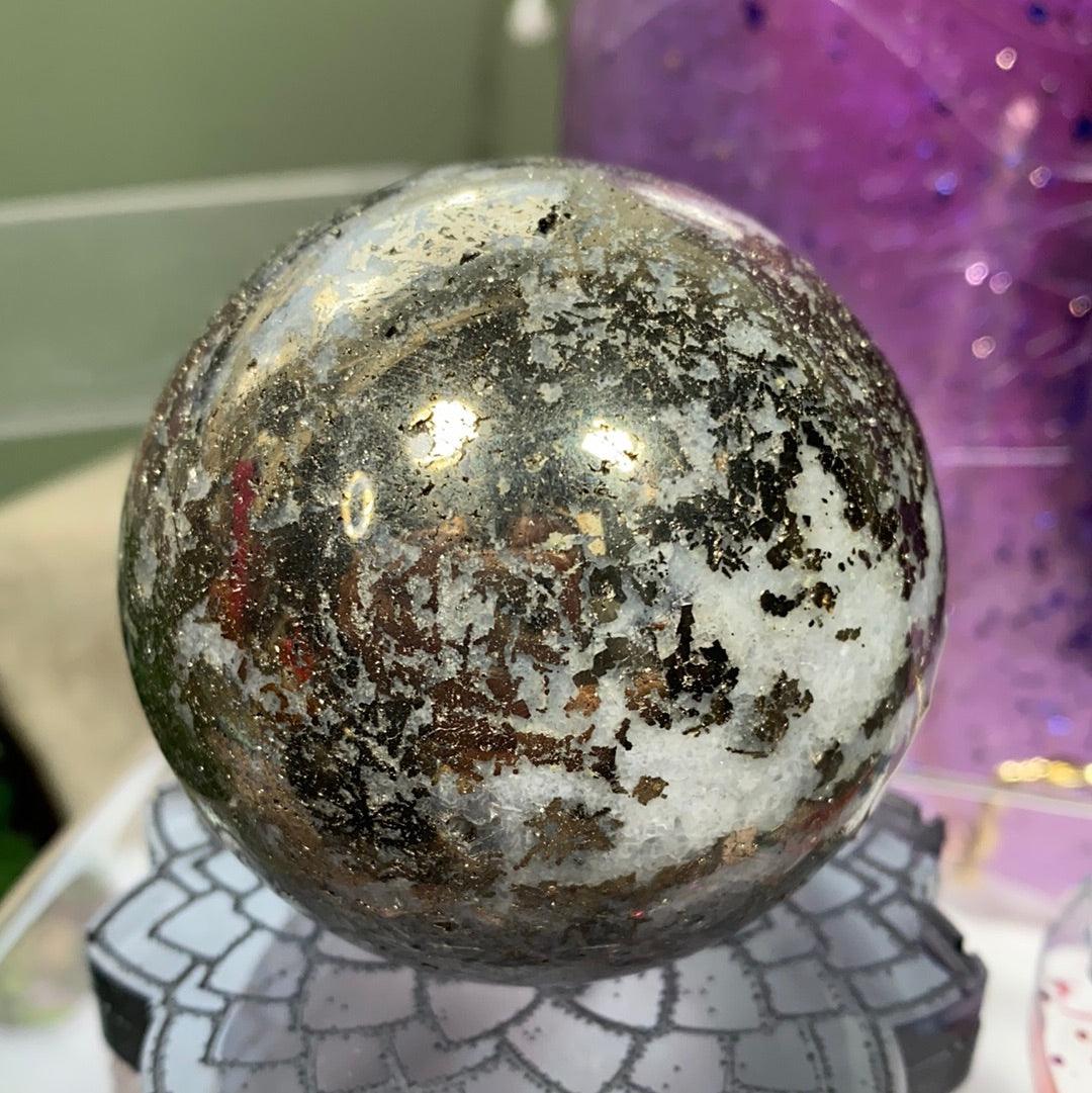 A close-up of The Crystalary's Pyrite in Quartz Sphere, showcasing a polished, spherical stone with a mixture of quartz, pyrite, and black and white speckles. The sphere is elegantly perched on a decorative, lotus-shaped stand against a purple, glittery backdrop that is beautifully out-of-focus.