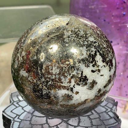 A Pyrite in Quartz Sphere from The Crystalary rests elegantly on a geometric-patterned display stand. The sphere boasts a blend of black, white, and gray hues, with reflective surfaces and sparkling inclusions of Pyrite. In the slightly blurred background, a purple object is visible.