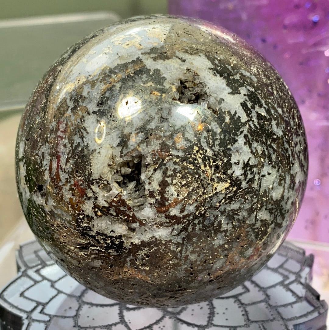 A polished sphere of Pyrite in Quartz from The Crystalary, exhibiting a metallic sheen with reflective silver and dark patches, sits atop a decorative stand. A druzy pocket adds texture to its surface. The background features a purple bokeh effect, creating a colorful, blurred ambiance.