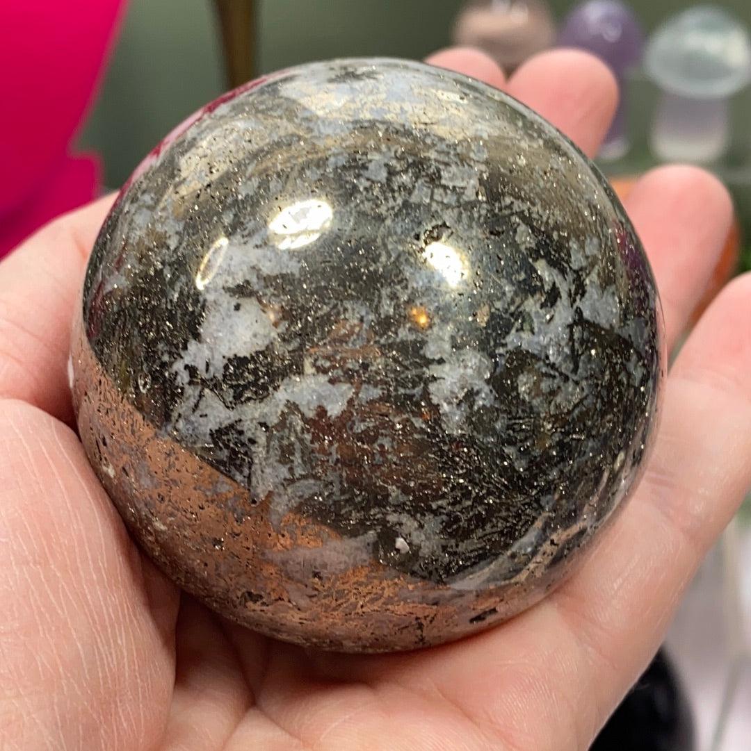 A hand holding a Pyrite in Quartz Sphere by The Crystalary, featuring a polished, spherical stone with a mix of dark and light patches that hint at quartz. The stone has a glossy surface adorned with an intricate pattern of streaks and specks in various colors. In the background, there is a pink object and blurred shapes.