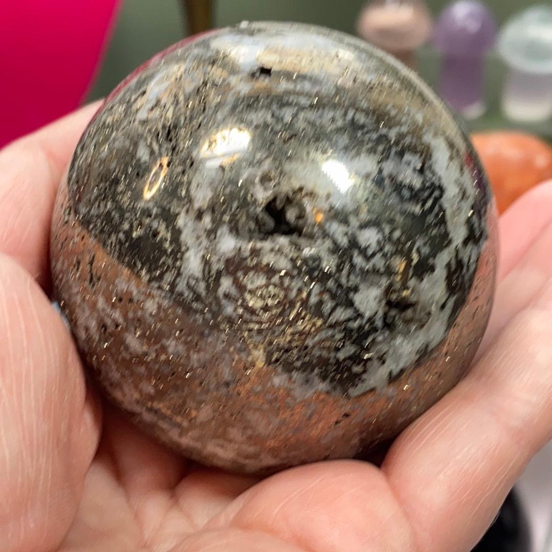 A hand is holding The Crystalary's Pyrite in Quartz Sphere, a smooth, polished ball with a marbled pattern. This sphere showcases a blend of earthy colors with shades of black, grey, white, and brown. It features gleaming streaks reminiscent of pyrite and hints of quartz that add to its reflective charm.